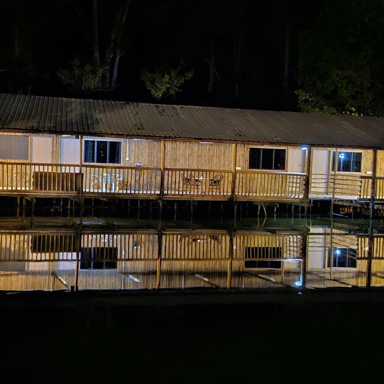 Night view of a long bamboo building on stilts reflected in calm dark water.