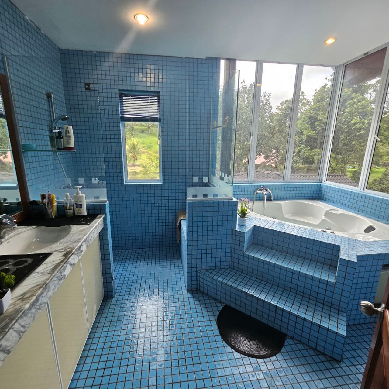 Bright blue tiled bathroom featuring a large white bathtub and windows overlooking lush greenery.