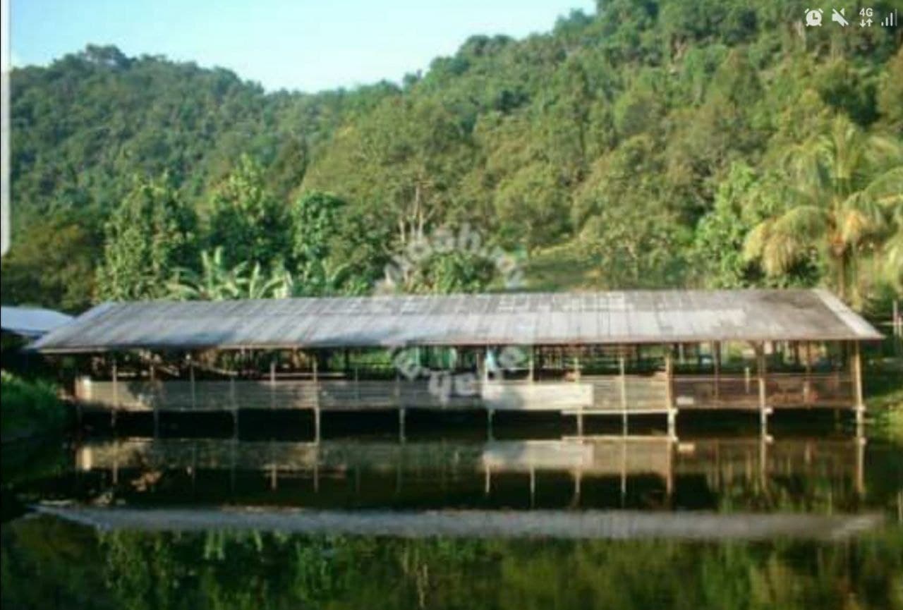 Long wooden shed on stilts over water, reflected, with lush green forested hills behind.