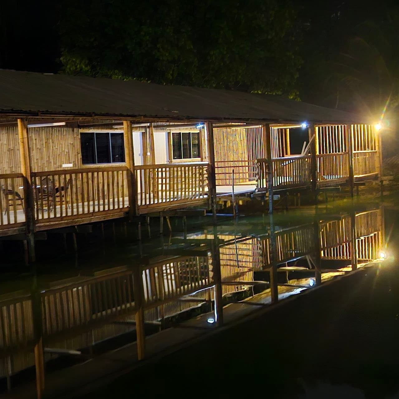 Warmly lit bamboo structure on stilts over water at night with a clear reflection.