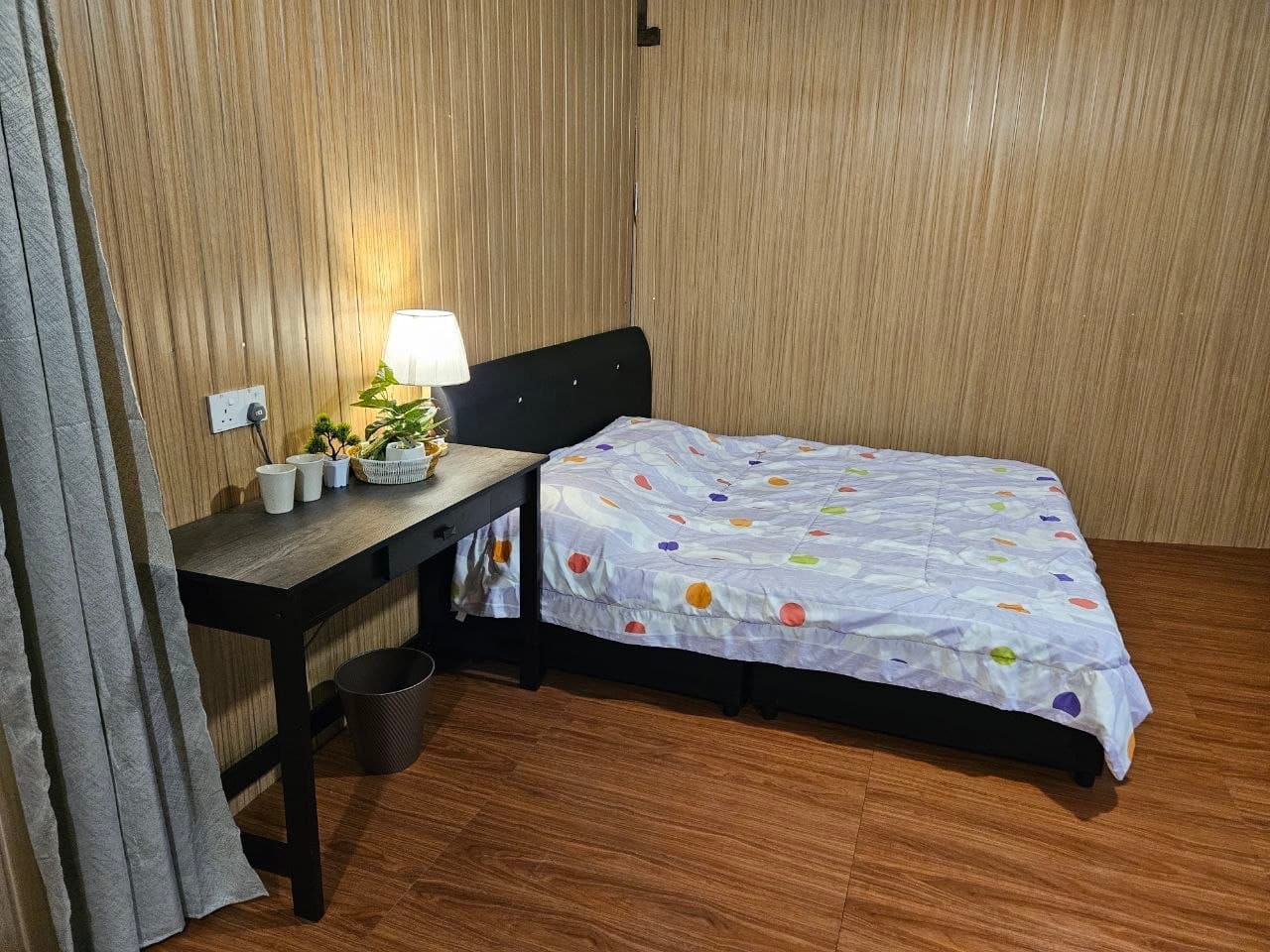 Cozy wood-paneled room with a black bed, patterned comforter, and a desk lamp.